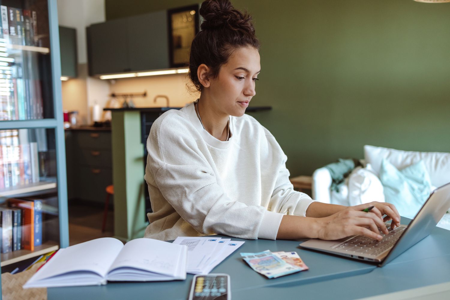 A woman sits at a table with a laptop and calculator, reviewing her budget 