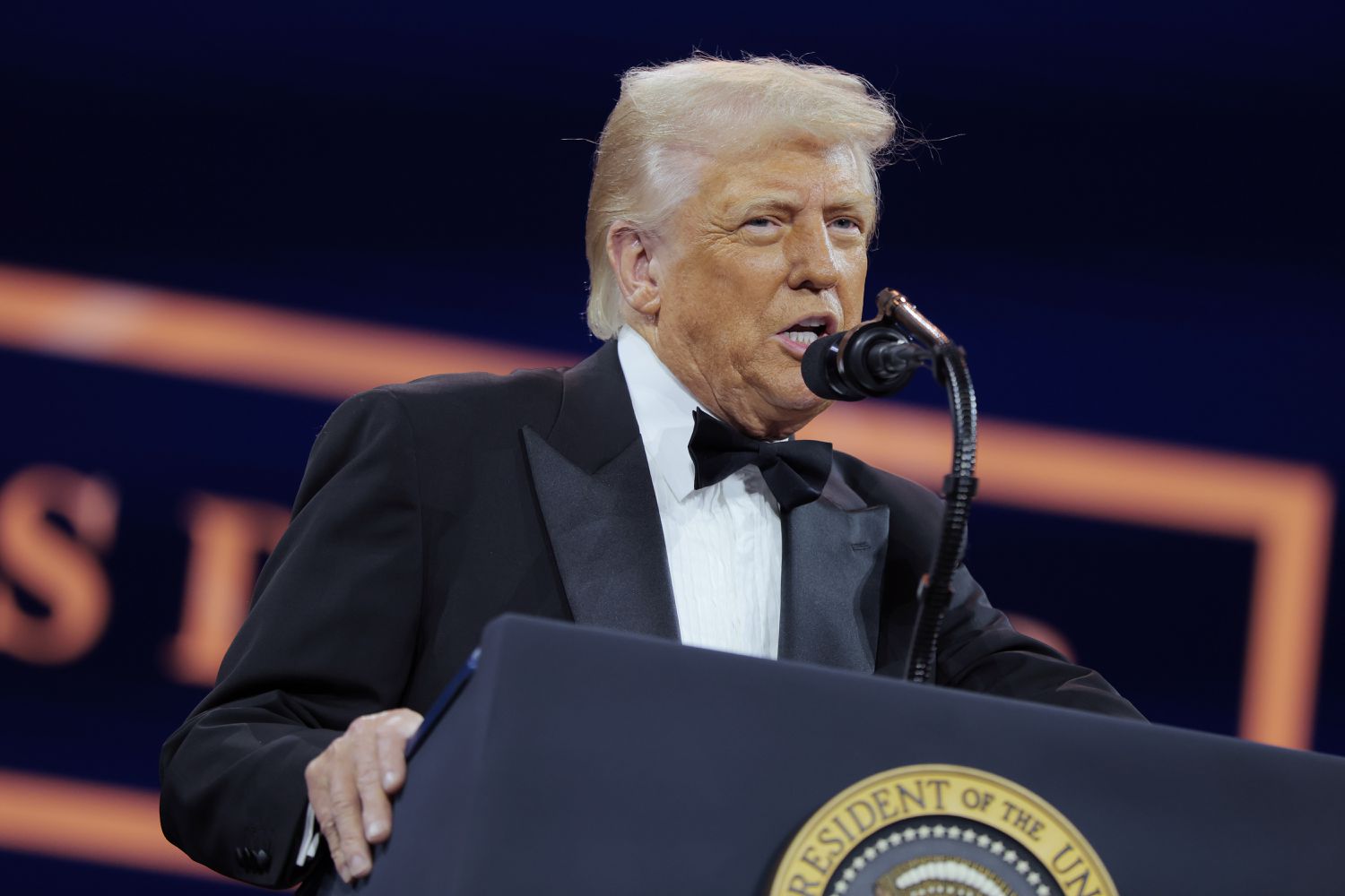 Donald Trump speaks in a black tuxedo in front of a podium and microphone.
