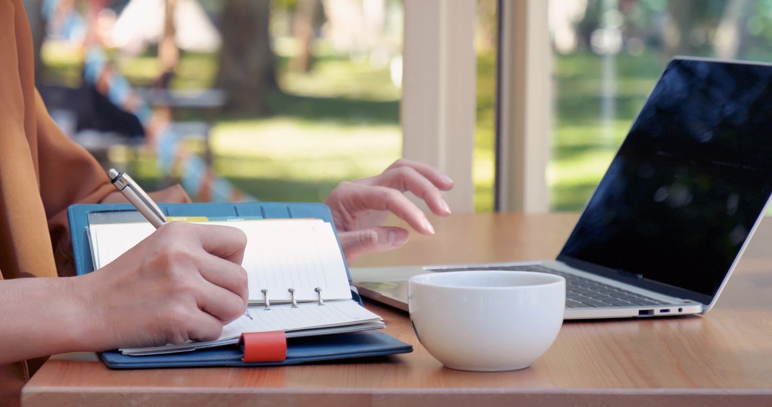 Hands of woman writing in a note pad on a desk with an open laptop and cup also on the desk.