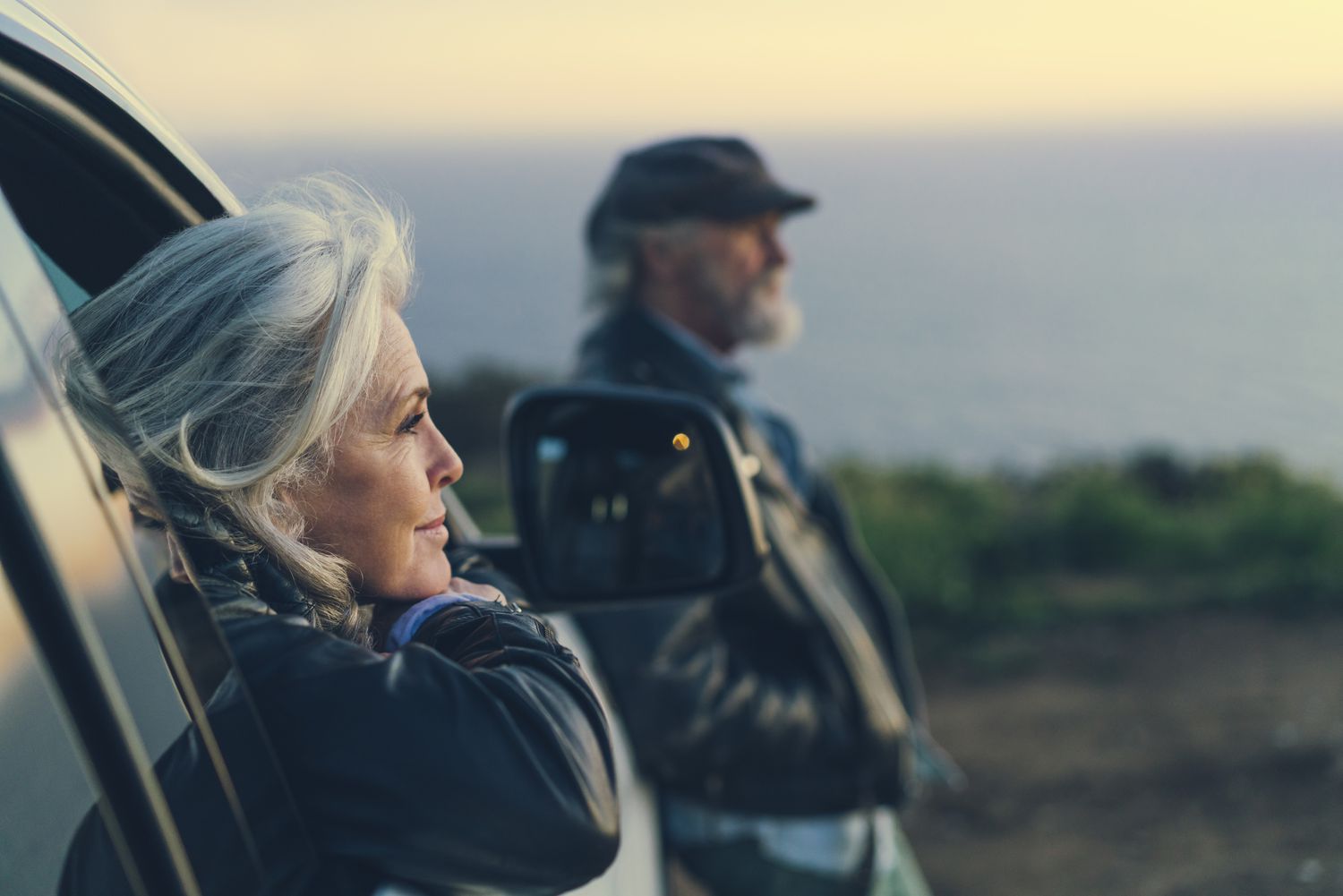 An older woman leans on a car door facing a sunset, while an older man leans against the ood