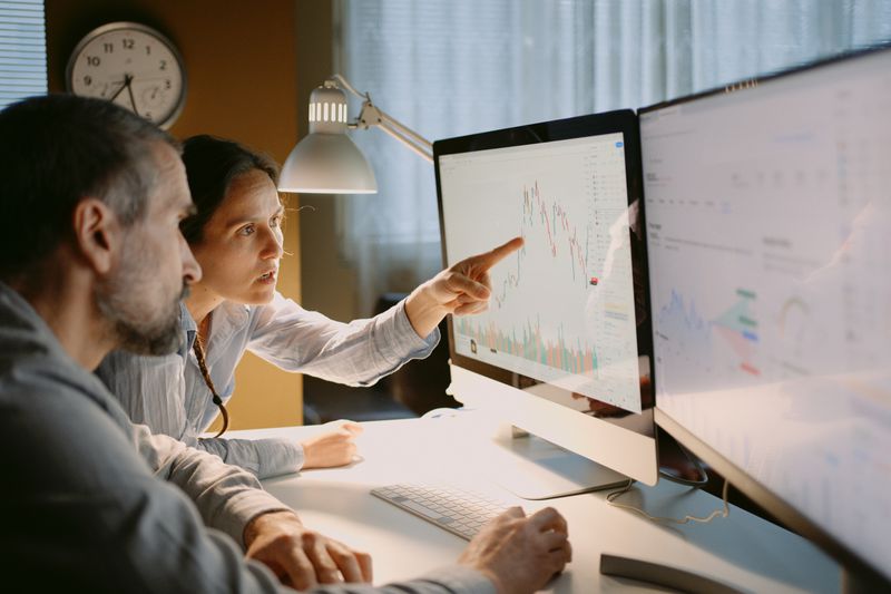 Two traders work at a dest with monitors showing stock data to check trading positions and speculate on market direction.