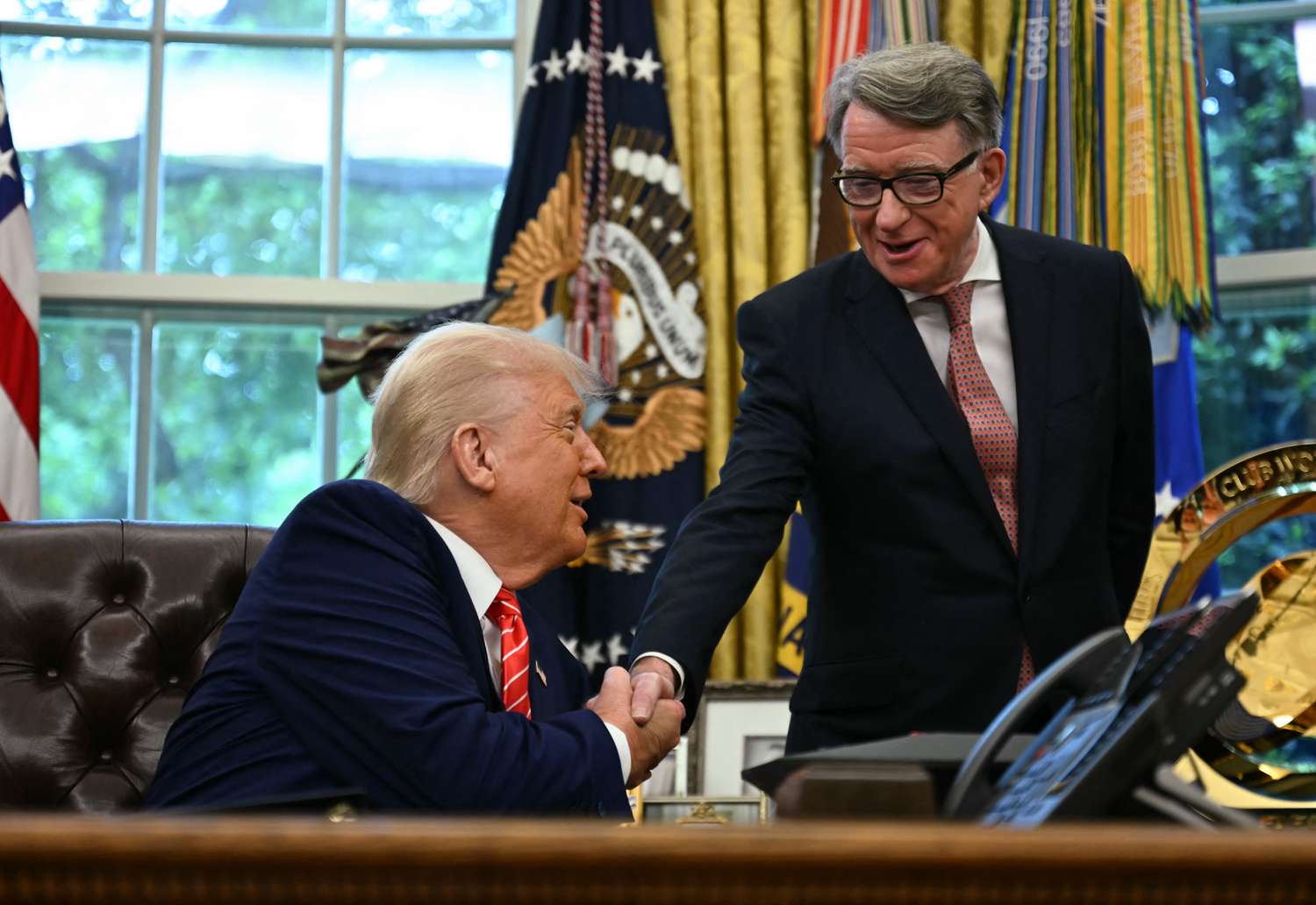 Donald Trump shakes hands with Peter Mandelson. Trump is sitting behind a desk in the Oval Office and Mandelson is standing to his left. 