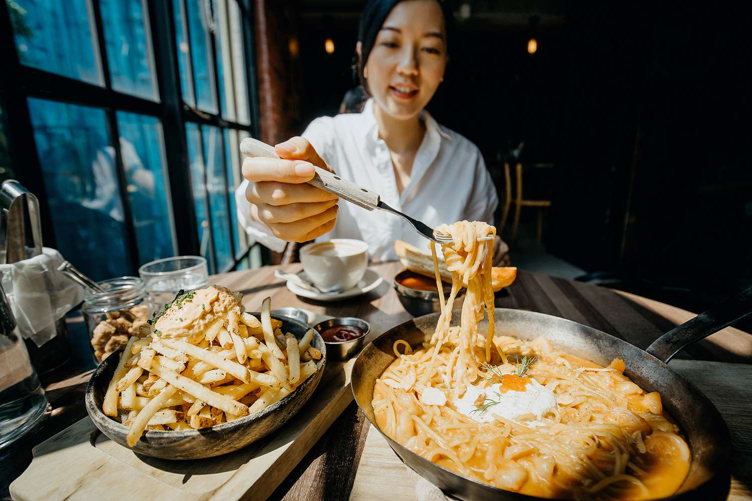 Young Asian Woman Sitting at a Table by the Window Enjoying the Warmth of Sunlight and Having Meal Joyfully in a Restaurant