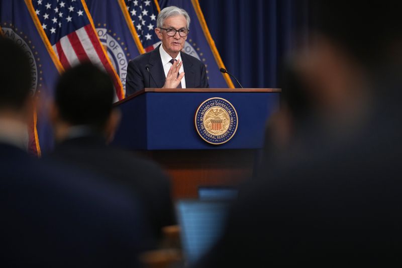 Jerome Powell stand in front of U.S. flags and Federal Reserve flags. He's in front of a podium, facing a crowd of reporters who are out of focus.