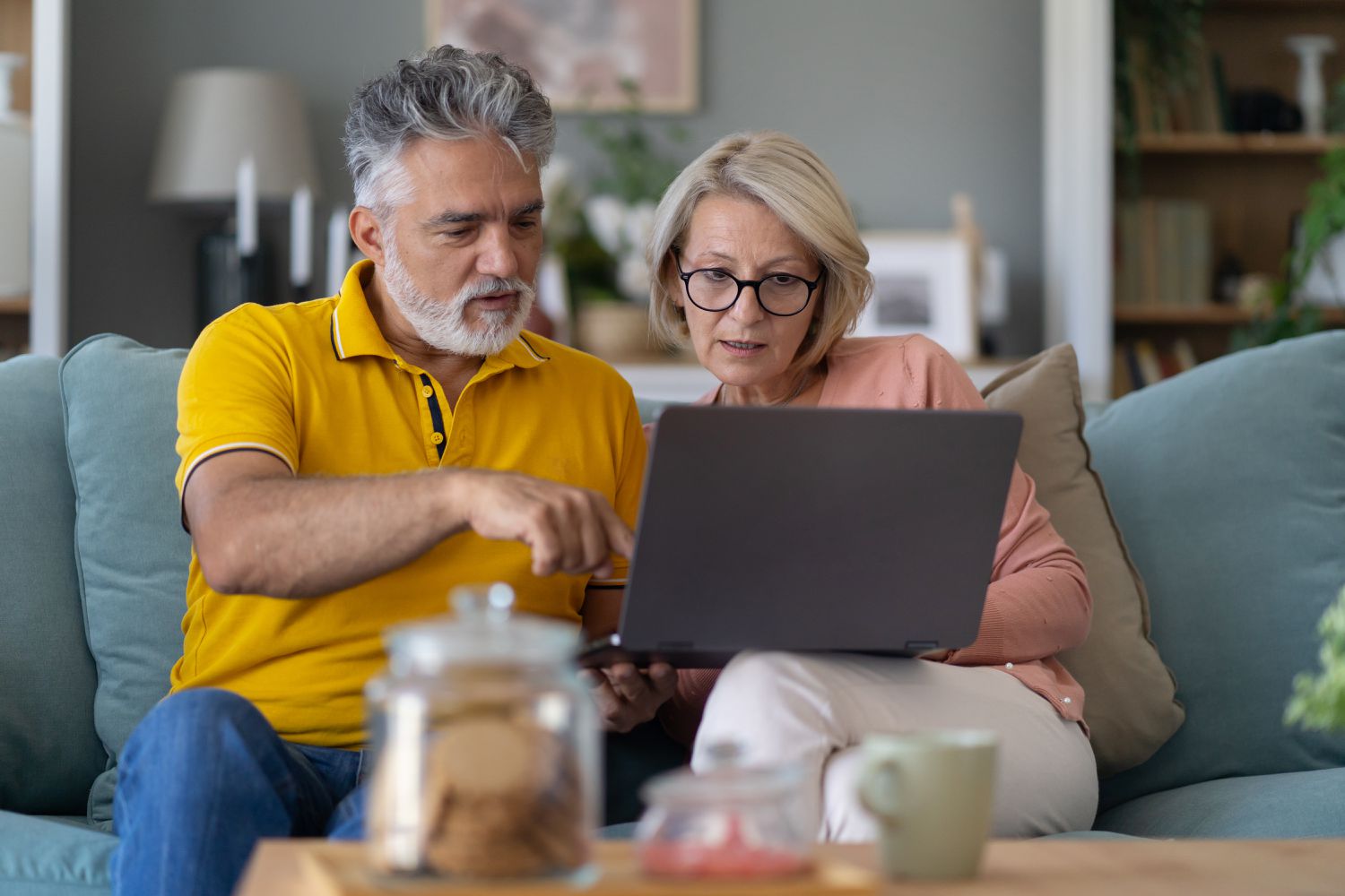 An adult couple sit together on a sofa using a laptop to review the interest and dividends earned on their retirement accounts.
