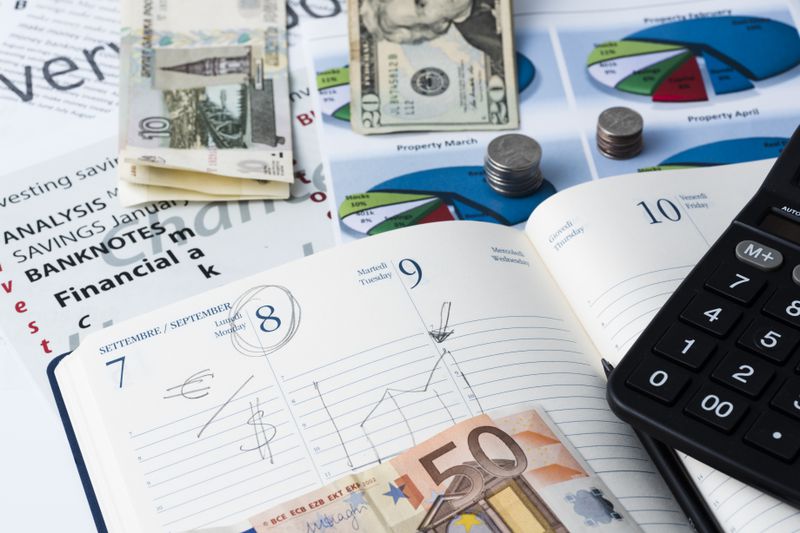 An appointment book shows a marked date with a note to exchange euros for U.S. dollars and laying around the book are stacks of various currencies.