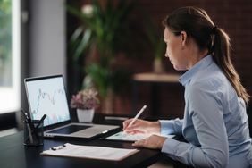 A woman in business attire sits at a desk and writes on a tablet while looking at a laptop displaying a financial chart.