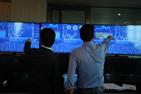 Two people pointing at screens with stock information while standing.