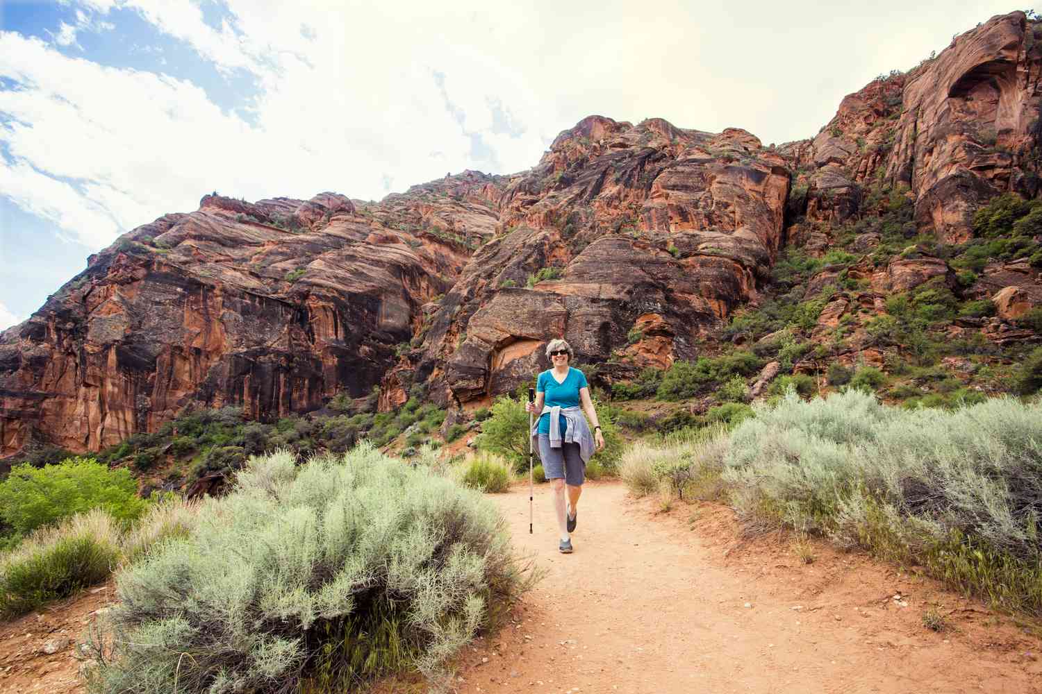 A retired woman is enjoying a walk along a scenic trail with vibrant red rock cliffs in the background.