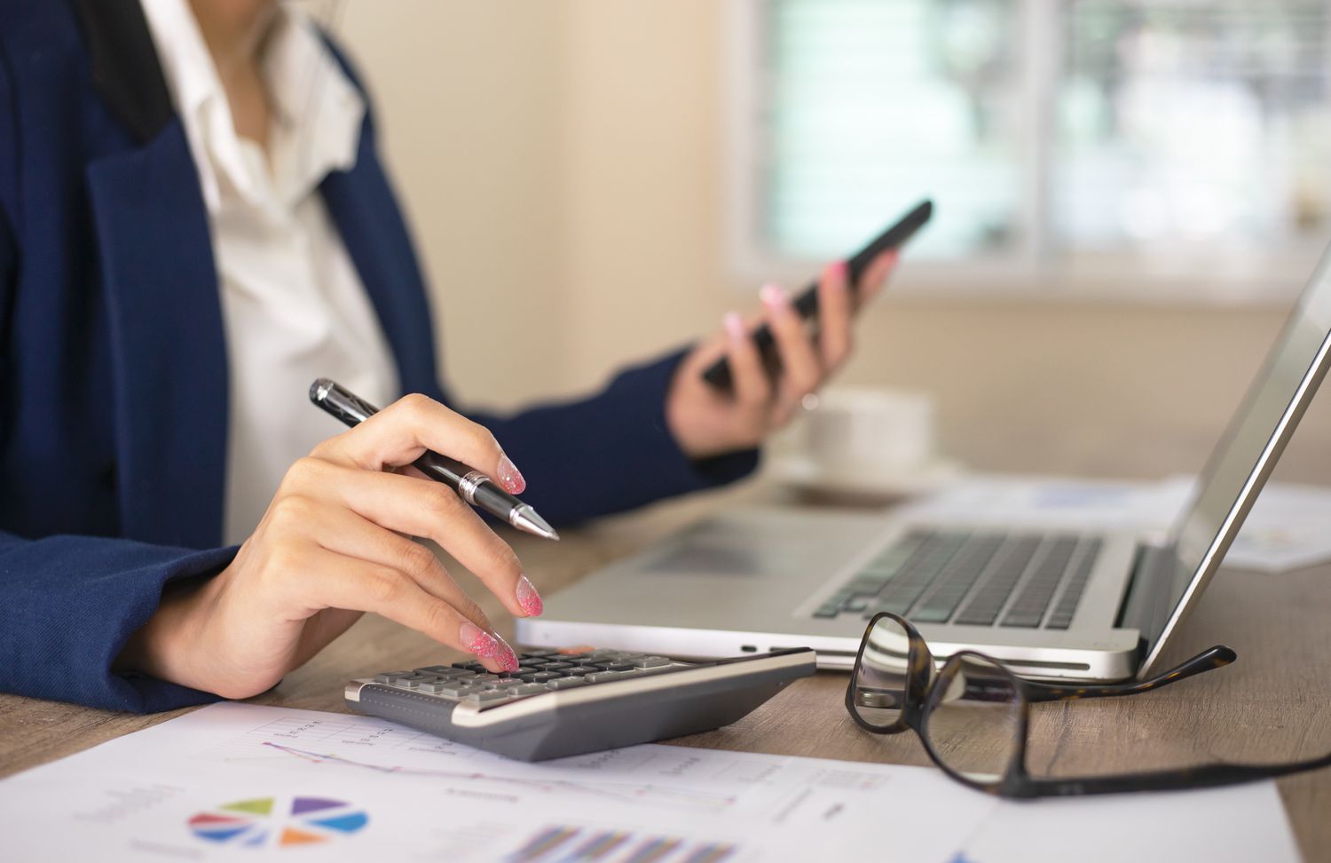 Businesswoman working in office typing on calculator and holding phone in hand with computer and financial sheets on desk
