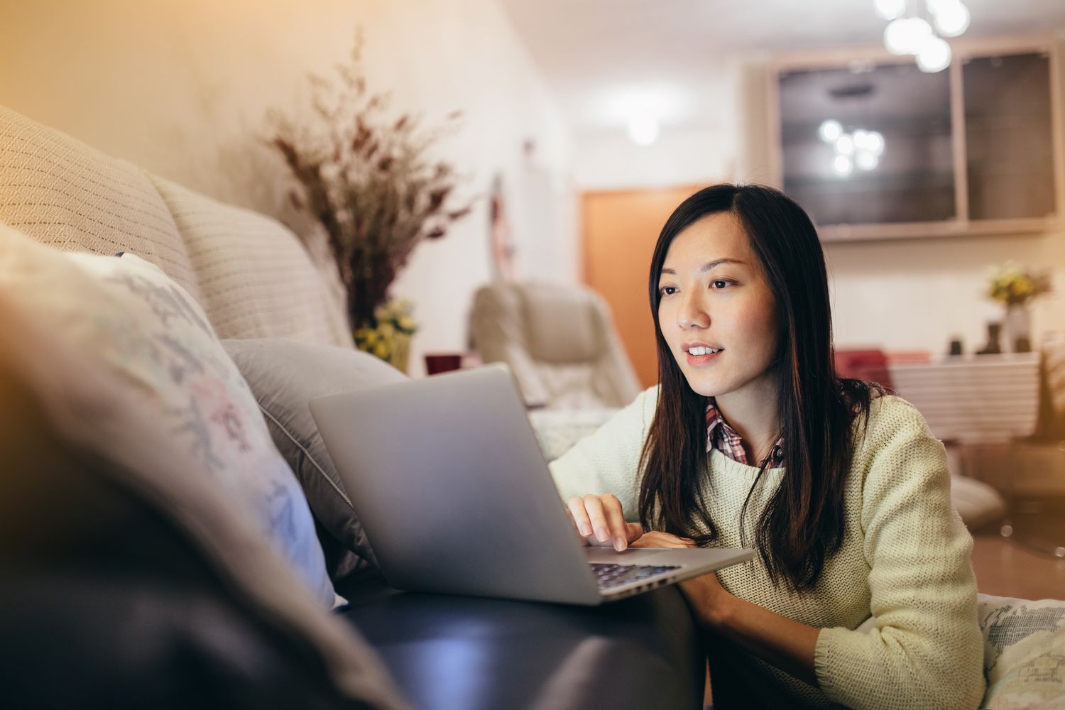 A woman wearing a white sweater sits on the ground facing a sofa looking at a laptop that is on the sofa