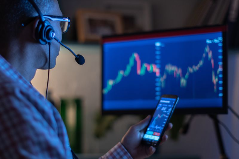 active investor at a computer screen, holding a phone, both showing stock charts. The trader is wearin a headset and sitting in the dark. 