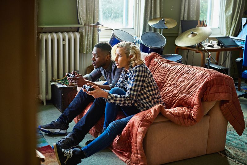 Two young adults playing console video games sitting on a couch in a small apartment.