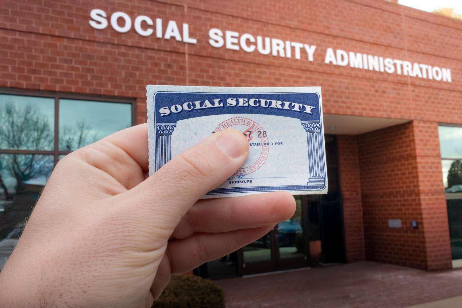 A hand holding a Social Security card, in front of the Social Security Administration building