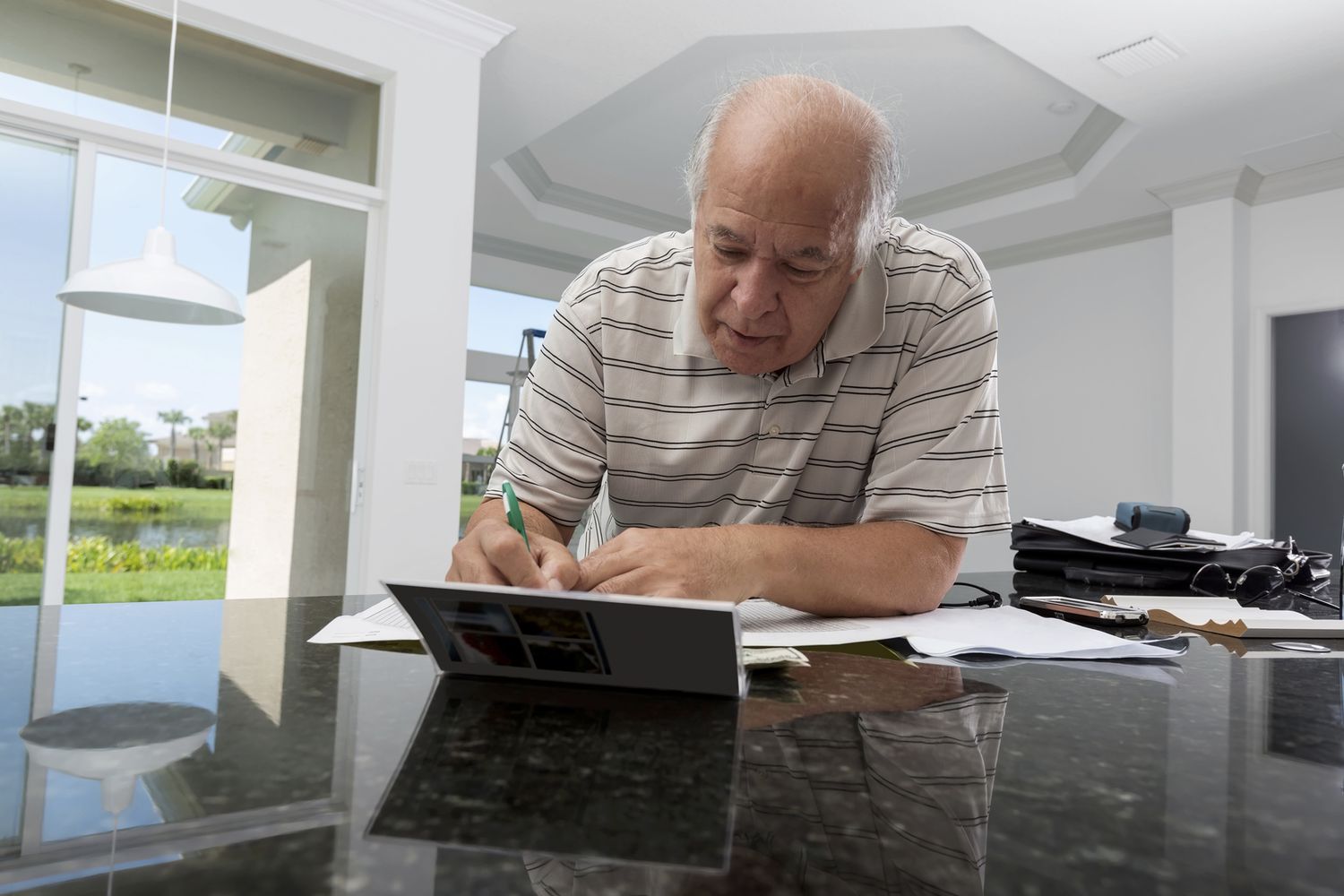 Older man at kitchen counter with laptop writing on a pad