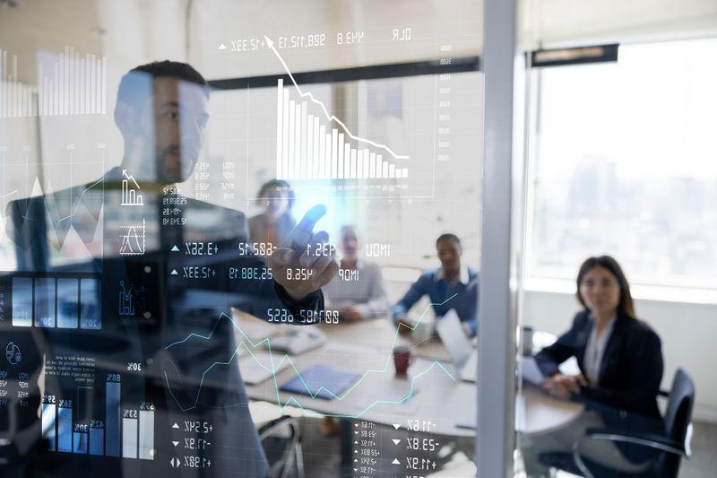 A man in a suit points to financial charts on a screen, presenting to people in a conference room