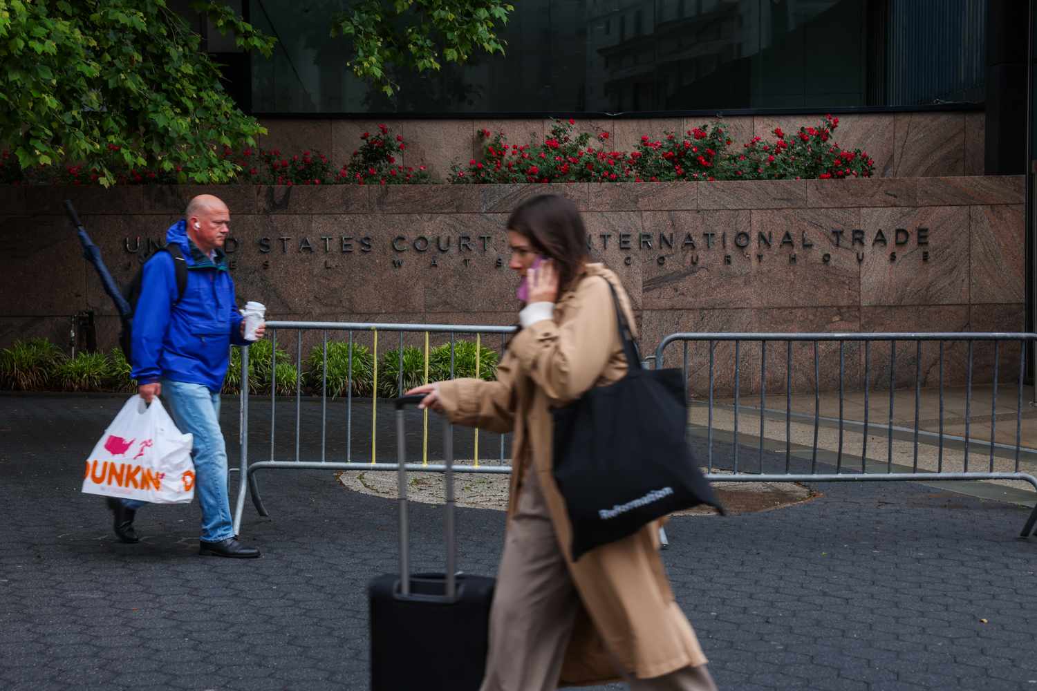 Two individuals walking near the United States Court International Trade building, one carrying a bag and coffee, the other pulling a suitcase