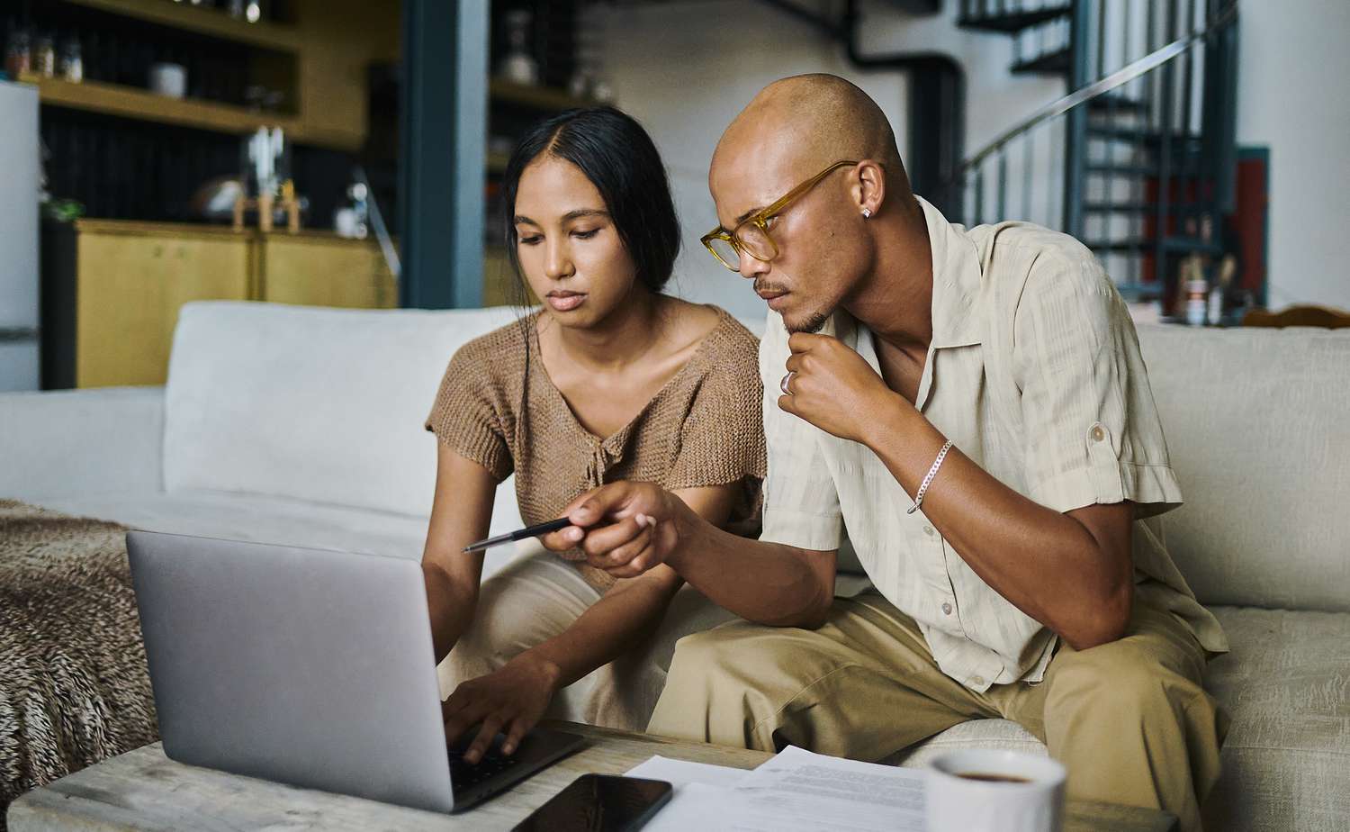 Two individuals sitting on a couch looking at a laptop, with papers and a cup on the table in front of them