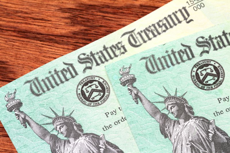 Two US Treasury checks of the type used to pay tax refunds as well as Medicare, Social Security and welfare benefits are partially visible on a wood desk background. Checks show the Treasury seal and image of the Statue of Liberty. 