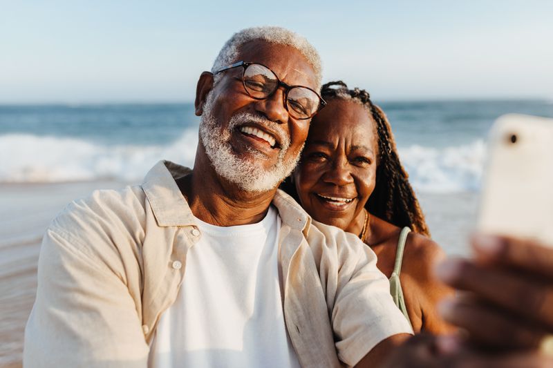 An older African American couple shares a joyful moment, taking a selfie together with the ocean backdrop during a serene beach walk.