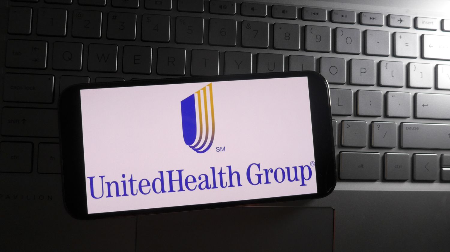 The blue and yellow UnitedHealth Group logo is seen on a phone screen, sitting on a laptop keyboard.