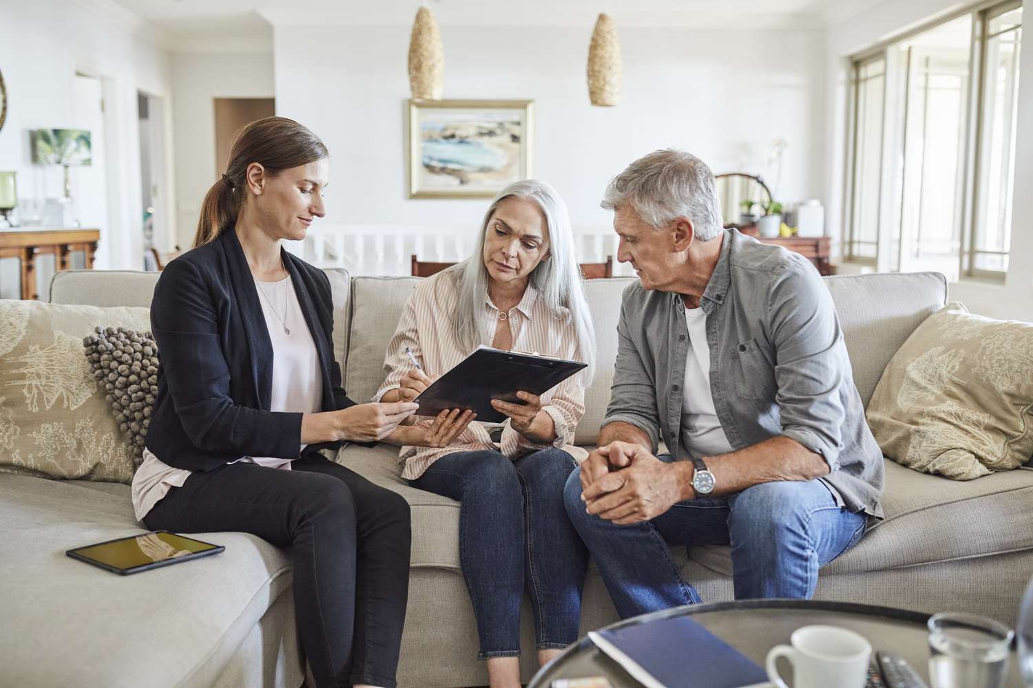 A woman shows a tablet to an older couple seated on a couch, having a conversation in a living room setting
